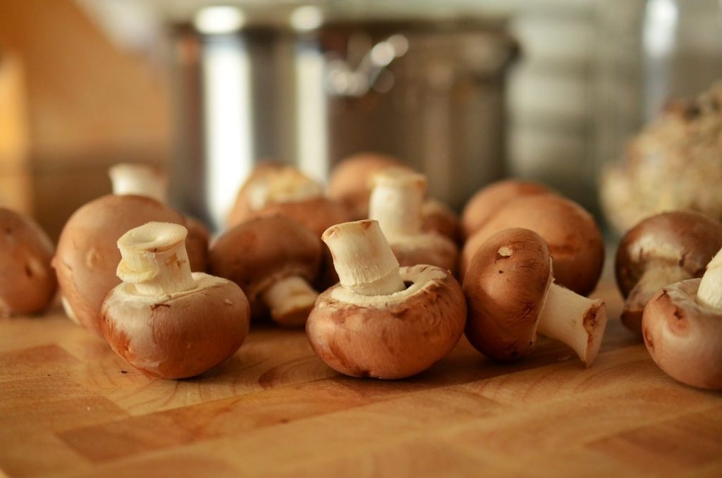 Fresh brown mushrooms scattered on a wooden cutting board, with a blurred stainless steel pot in the background, evoking a warm kitchen atmosphere.