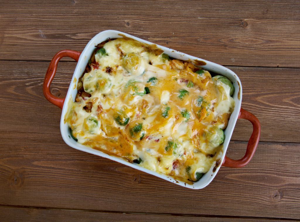 A baked casserole dish filled with chicken, vegetables, and melted cheese, served in a white dish with red handles on a wooden table.
