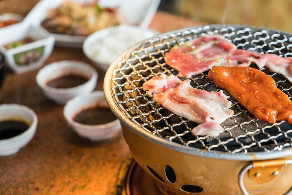 Slices of marinated pork grilling on a tabletop charcoal grill, with various dipping sauces and side dishes in the background.