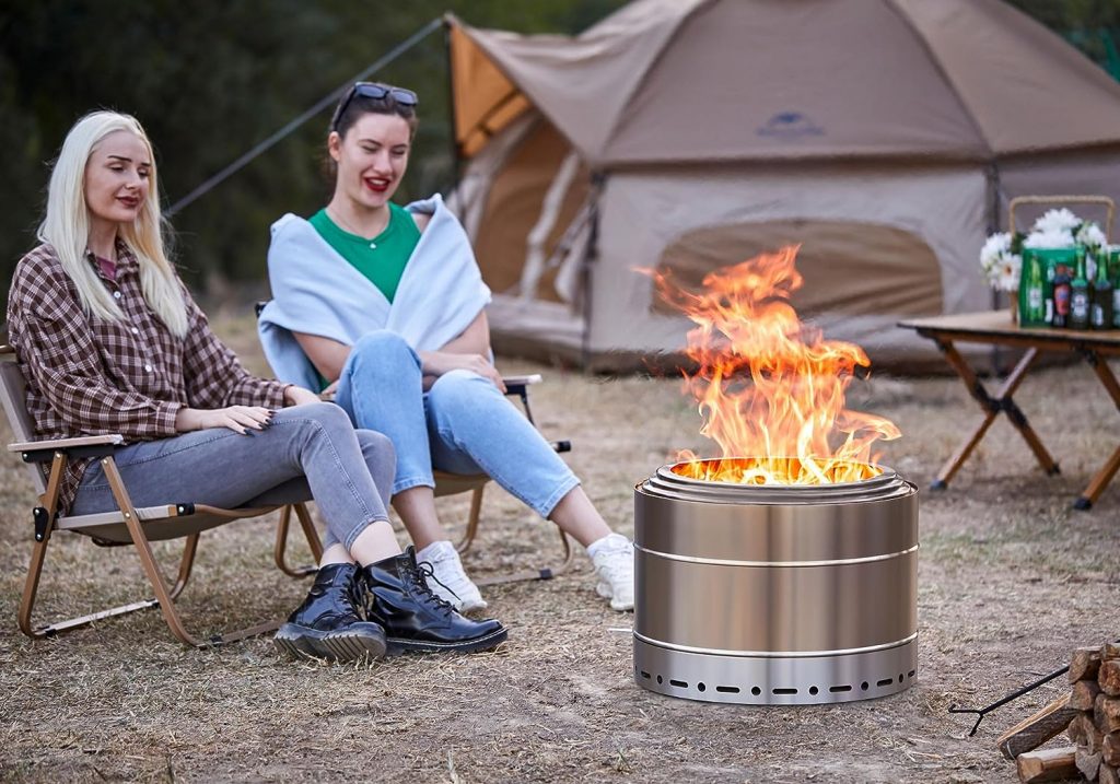 Two women sitting near a stainless-steel fire pit, enjoying the warmth, with a tent and camping gear in the background.